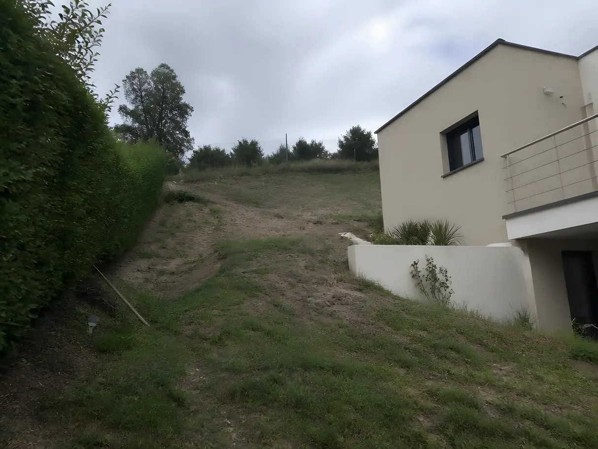 Talus raide d'herbe rase et de terre nue, raviné, contre le pignon d'une maison récente