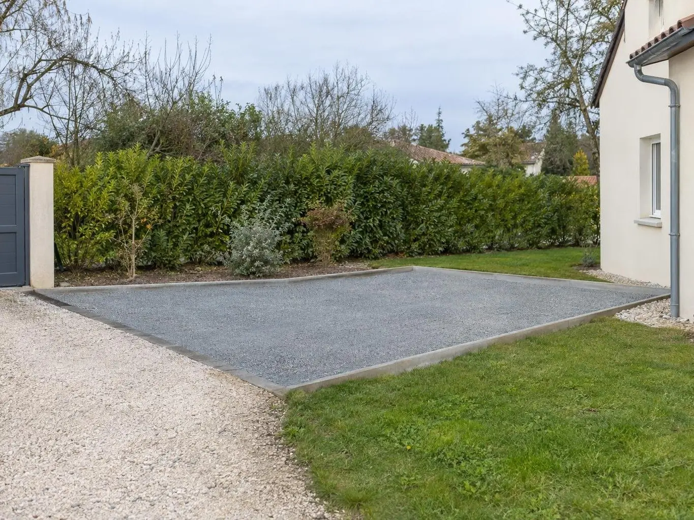 Plateforme rectangulaire de cailloux gris compactés tenue par des bordures béton, à côté du pavillon