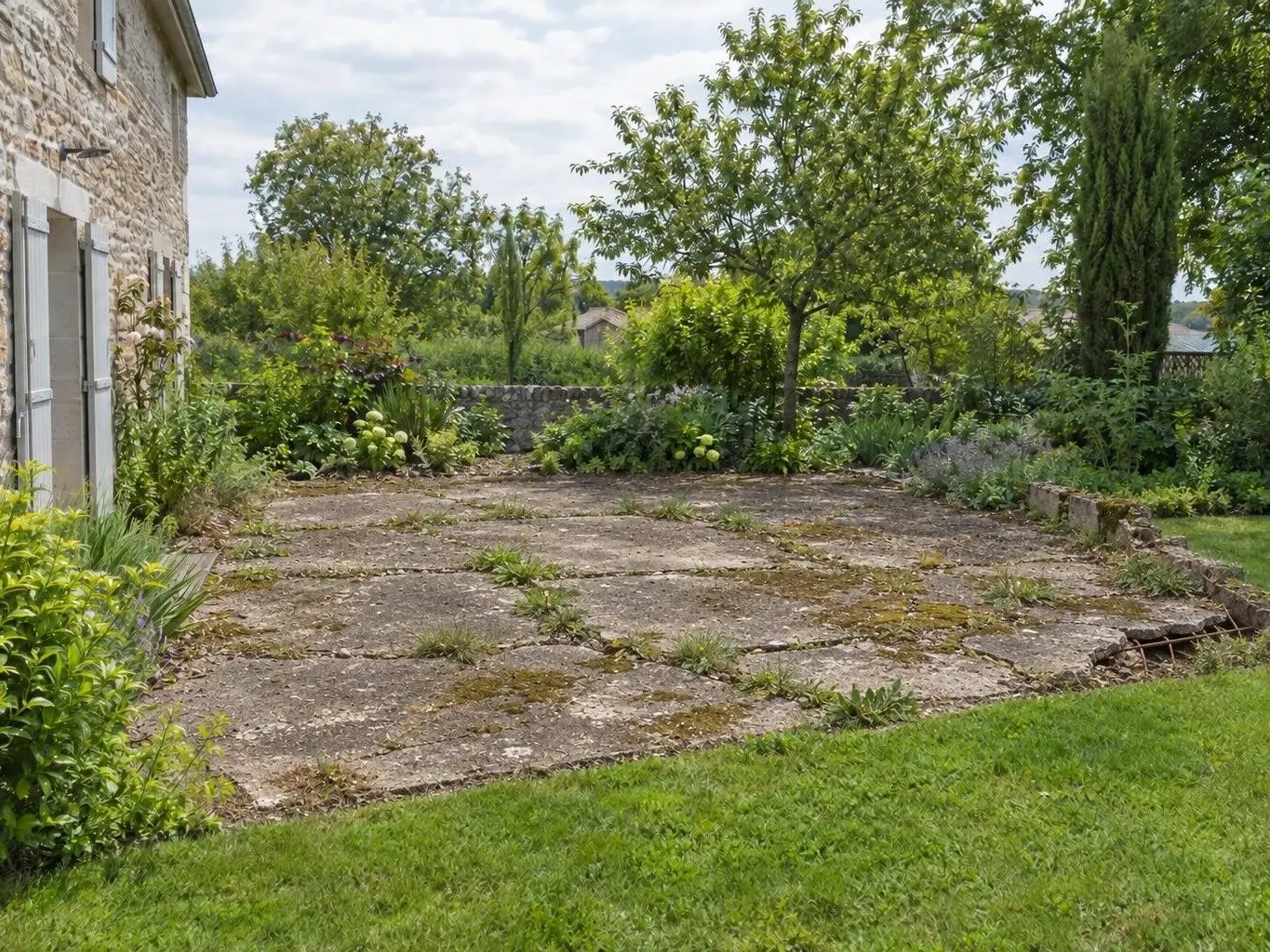 Vieille dalle de béton fissurée envahie de mousse dans le jardin d'une maison en pierre, muret arasé au bord