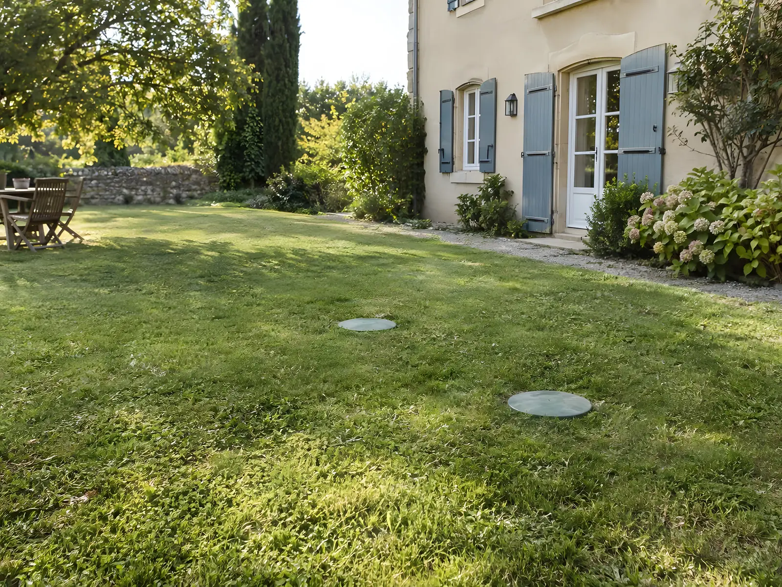 Pelouse verte et dense devant une maison aux volets bleus, deux petits couvercles ronds de cuve au ras de l'herbe