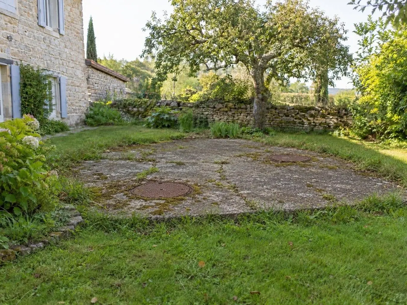 Vieille dalle de fosse en béton fissuré, deux tampons ronds rouillés, dans un jardin près d'une maison en pierre