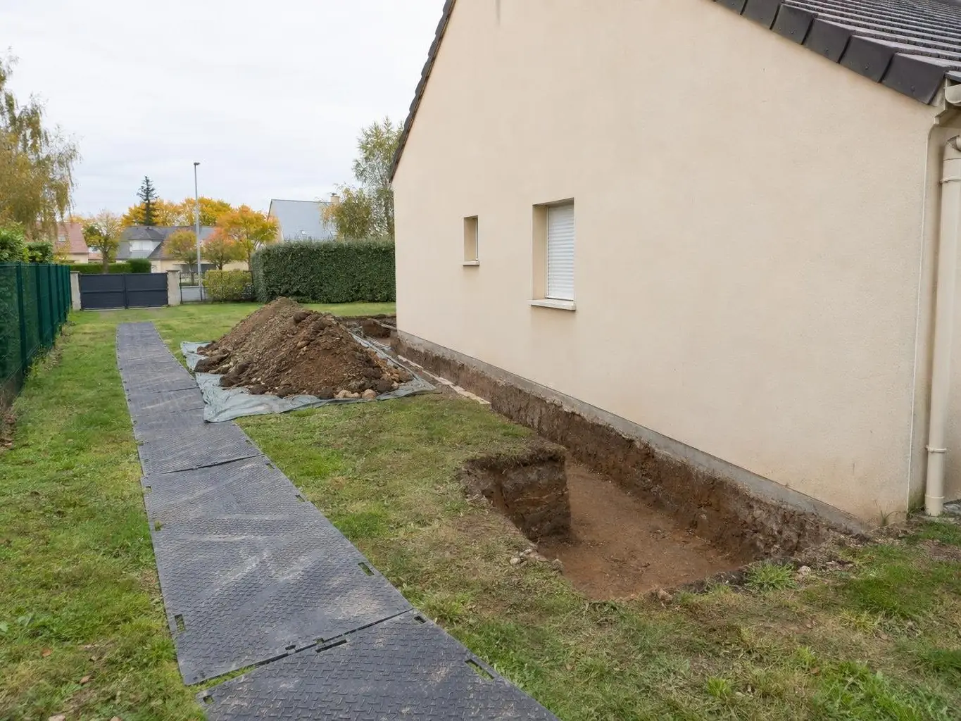 Fouille ouverte au pied du pignon d'un pavillon, plaques de roulage noires sur la pelouse, terre sur bâche