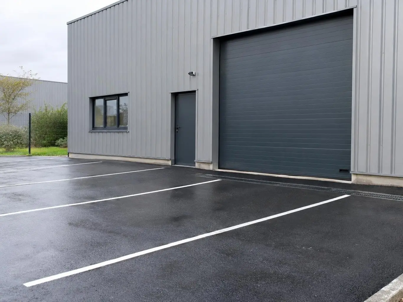 Parking en enrobé noir neuf aux places tracées en blanc devant un atelier en bardage métallique gris