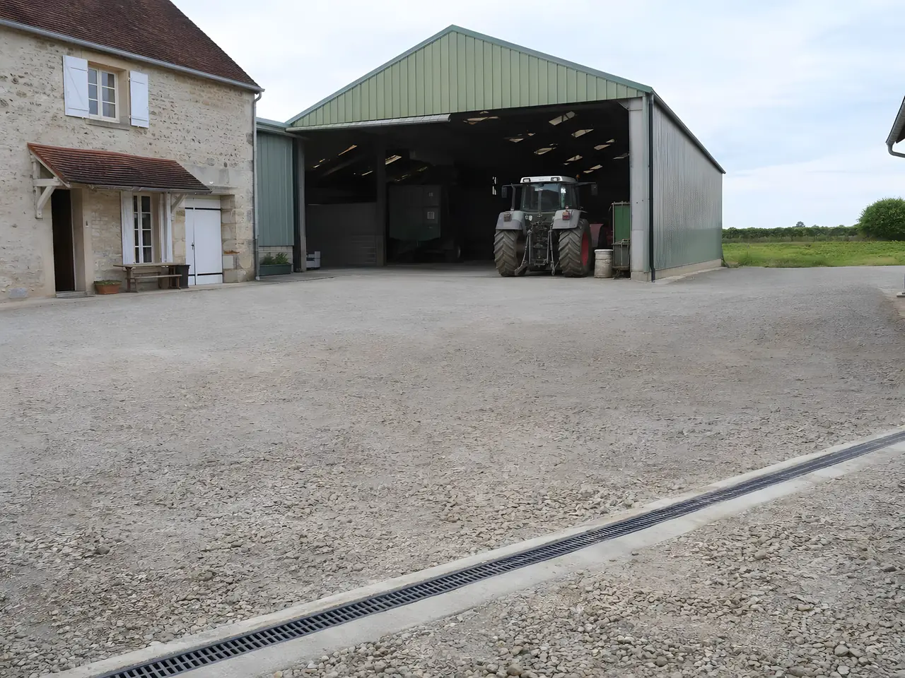 Grande cour de ferme en cailloux compactés devant un hangar vert abritant un tracteur, caniveau à grille au premier plan